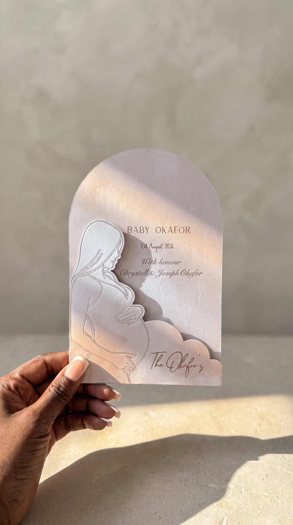 A hand holds a 3D embossed papercraft baby shower invitation card with a pregnant silhouette and embossed gold text, set against a textured beige wall with warm sunlight.