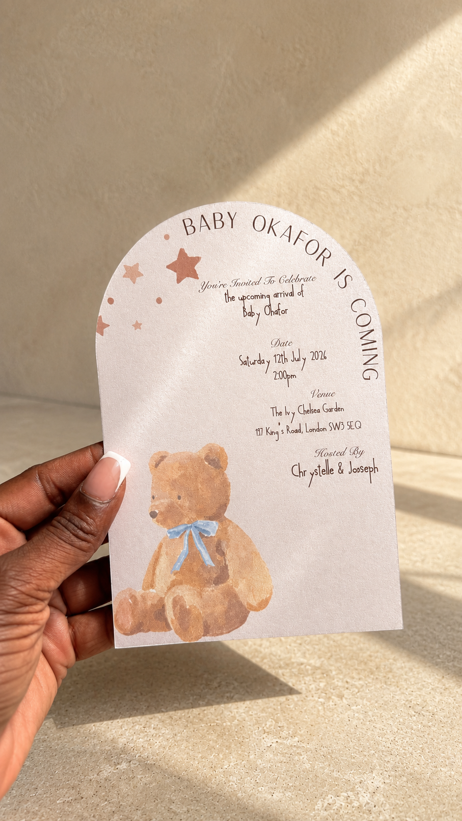 A hand holding a white arched baby shower invitation card featuring a watercolor teddy bear with a blue bow and the text 'BABY OKAFOR IS COMING'.