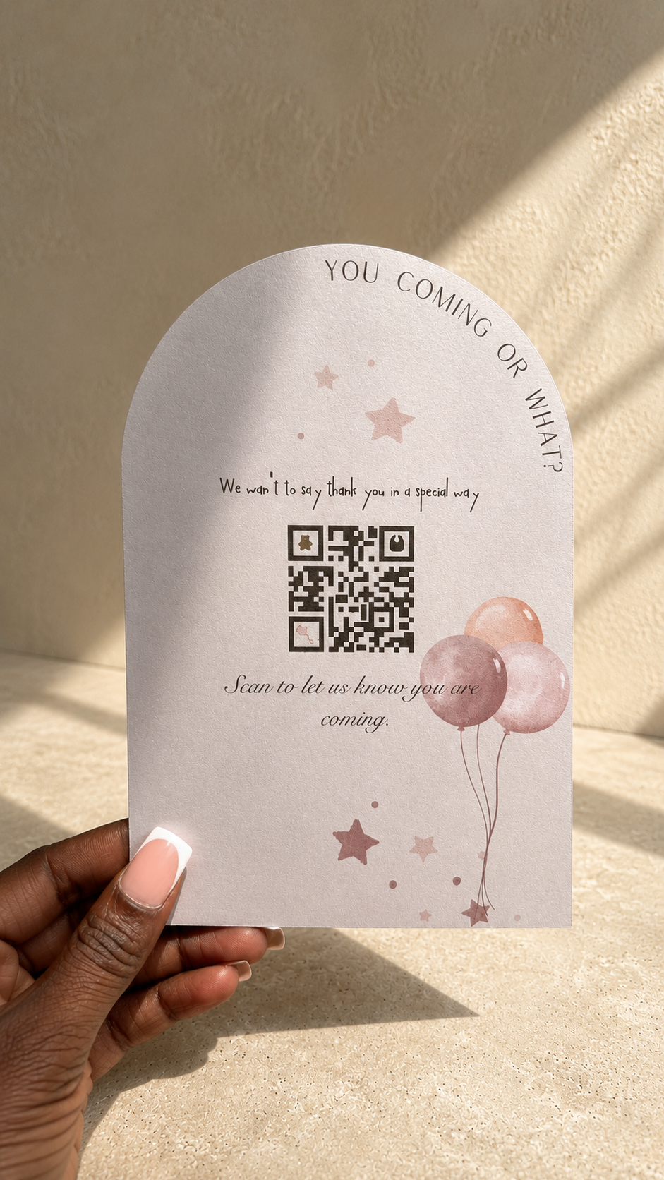 A hand with manicured nails holds a white arched card featuring a placeholder message, a QR code, and illustrations of pink balloons and stars against a sunlit beige wall.