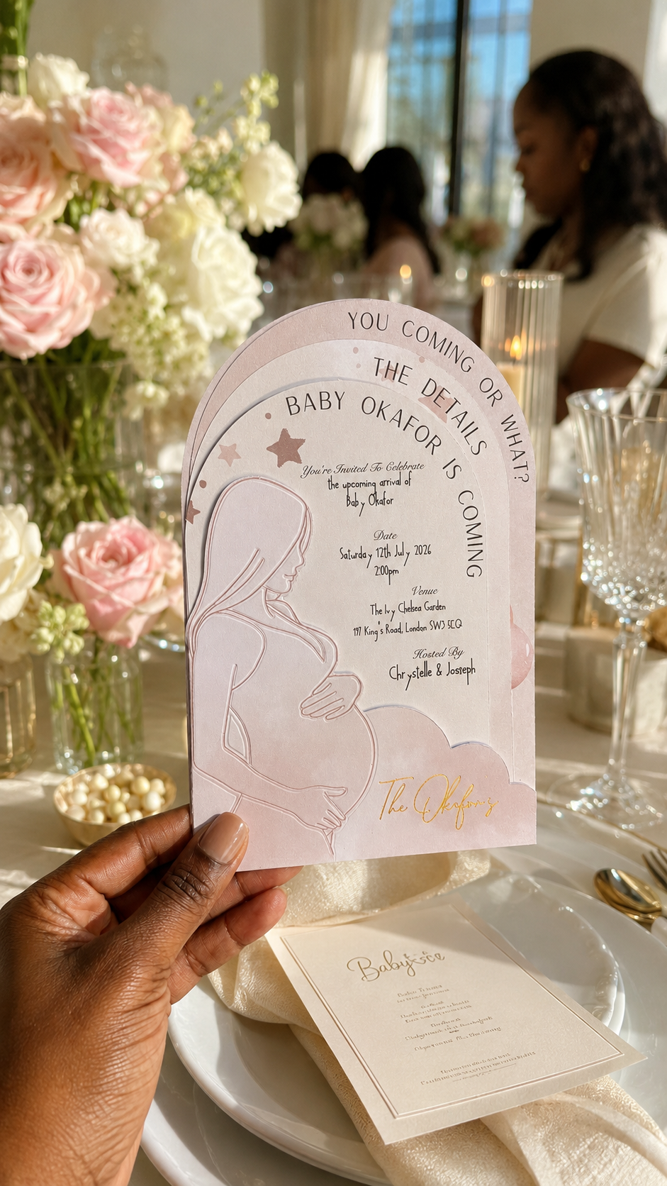 A hand holds a pink and white baby shower game card in a decorated event setting with flowers, plates, and glassware in the background.