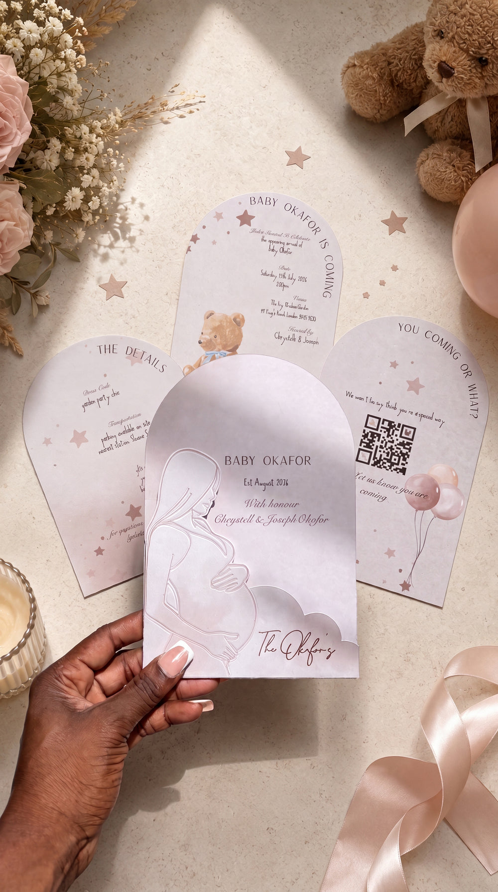 A hand holds a pastel pink arched baby shower invitation card with a line drawing of a pregnant figure, surrounded by matching invitation cards, dried flowers, a teddy bear, and a pink balloon on a light beige surface.