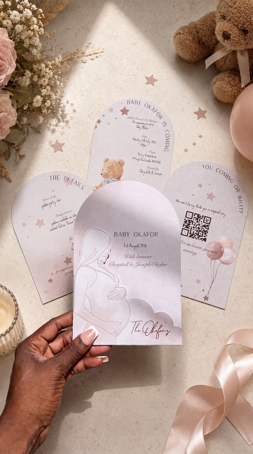 A hand holds a pastel pink arched baby shower invitation card with a line drawing of a pregnant figure, surrounded by matching invitation cards, dried flowers, a teddy bear, and a pink balloon on a light beige surface.